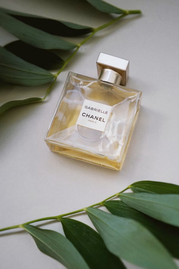 Gabrielle perfume bottle laid down on its side, logo facing up with green leaves around it.