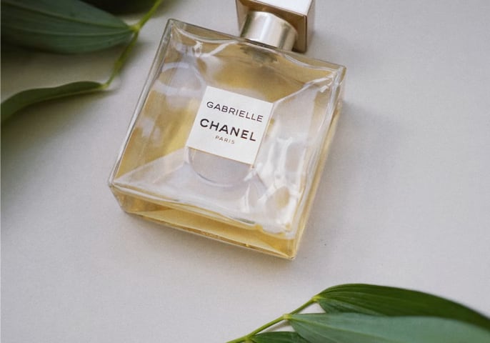 Gabrielle perfume bottle laid down on its side, logo facing up with green leaves around it.